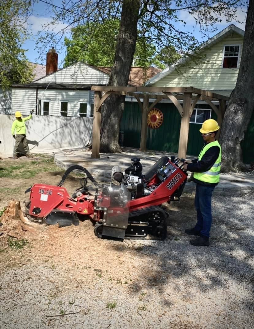 Two workers in hard hats beside a red forestry machine in a gravel driveway outside a house.