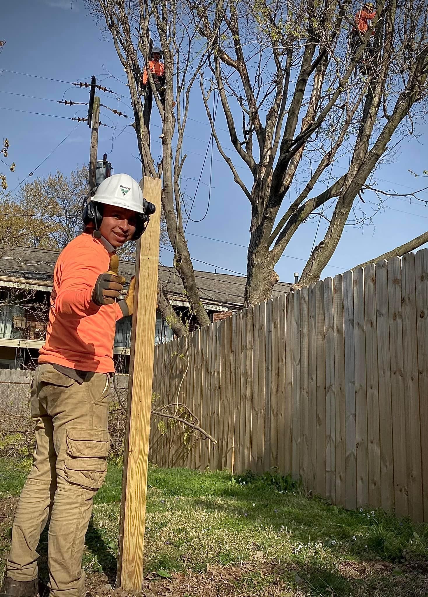 Worker in orange shirt and white hard hat standing by a wooden fence holding a tall post in a backyard.