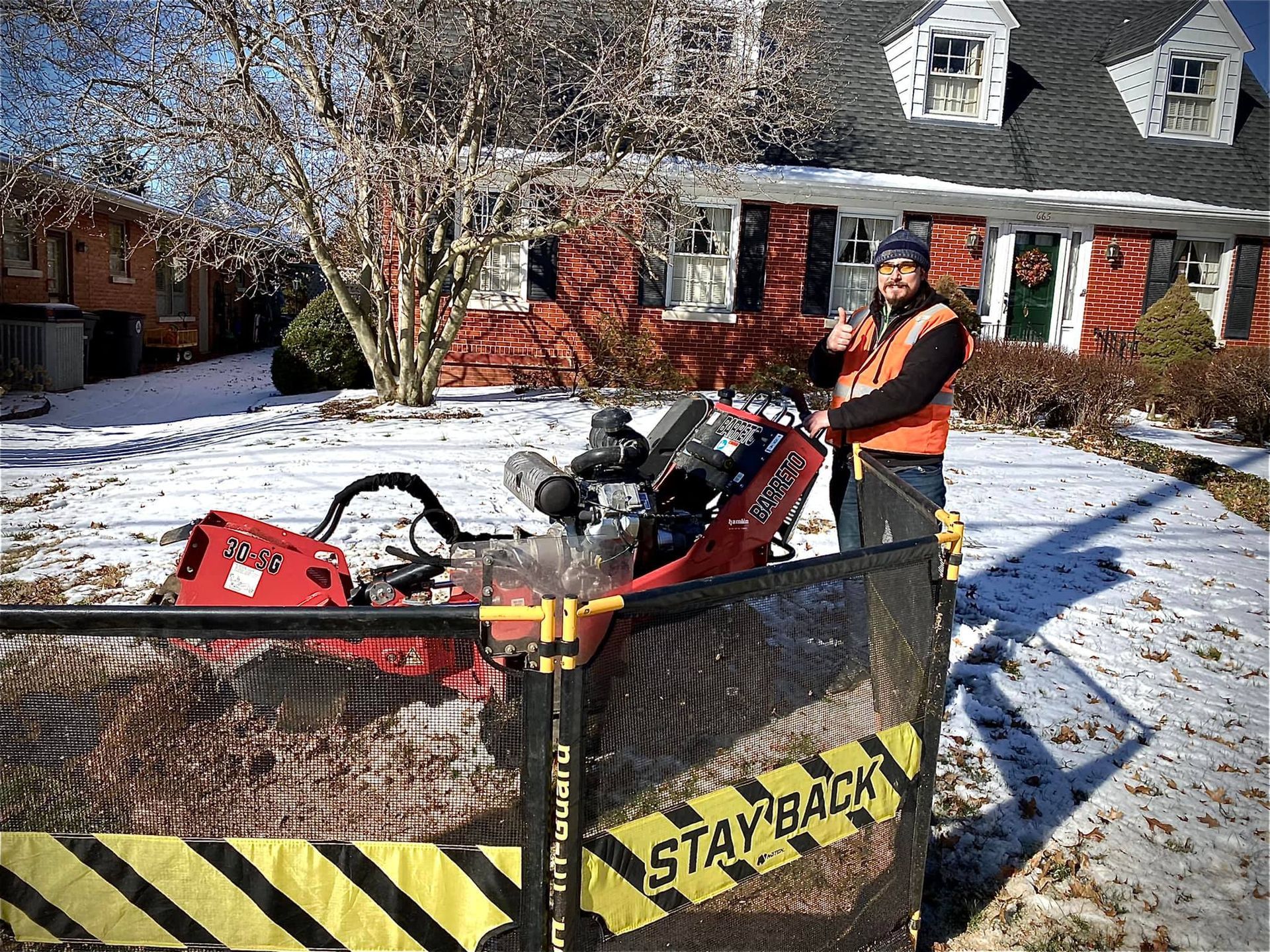 Worker in orange vest removing snow from a driveway beside a house, behind a yellow 