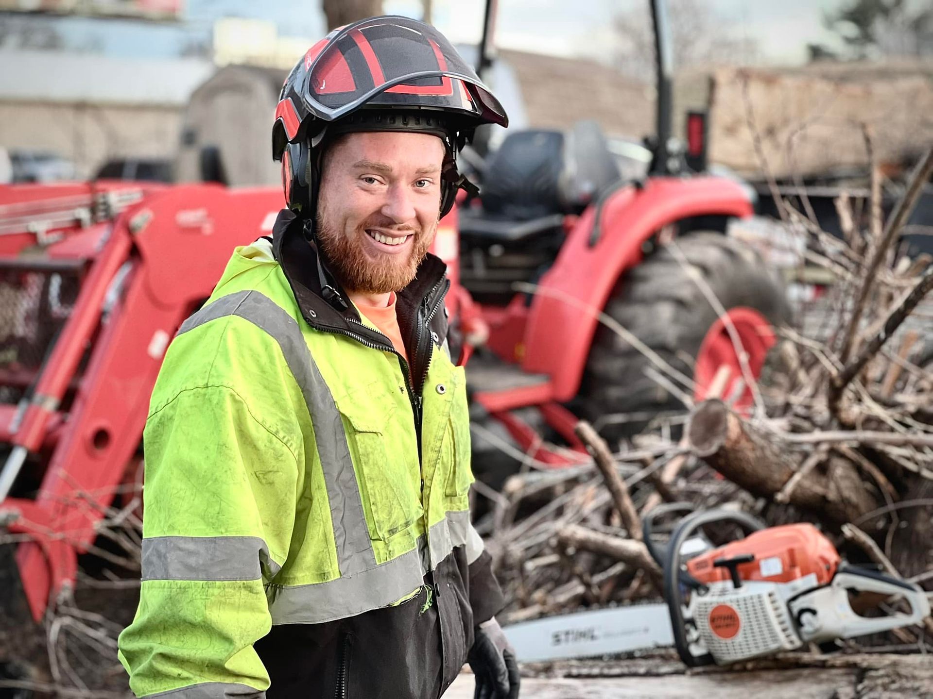 Worker in high-visibility jacket and hard hat smiling beside a chainsaw and tractor in a rural worksite