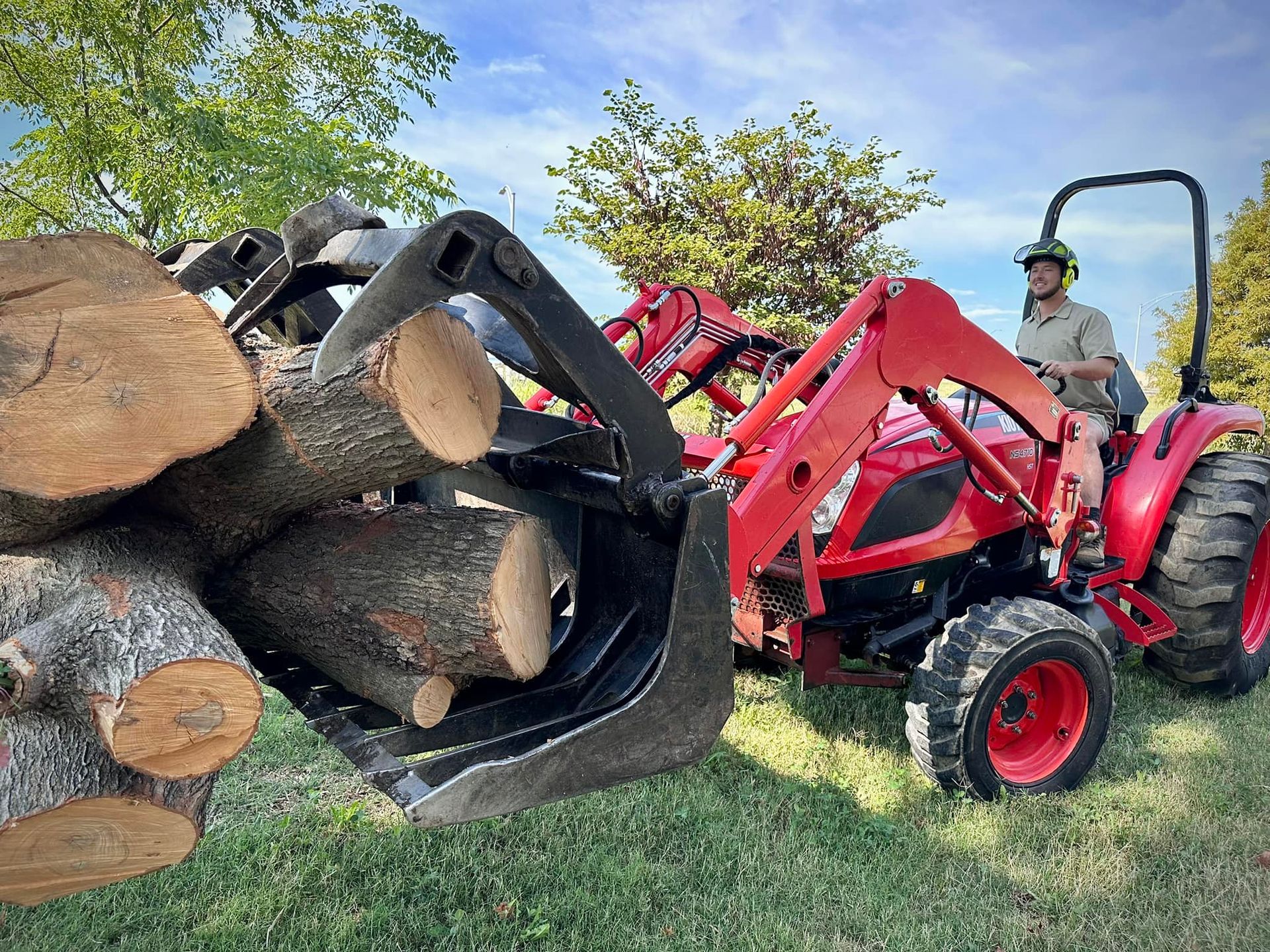 Tractor with front loader lifting cut logs in a grassy field, operator seated beside the pile