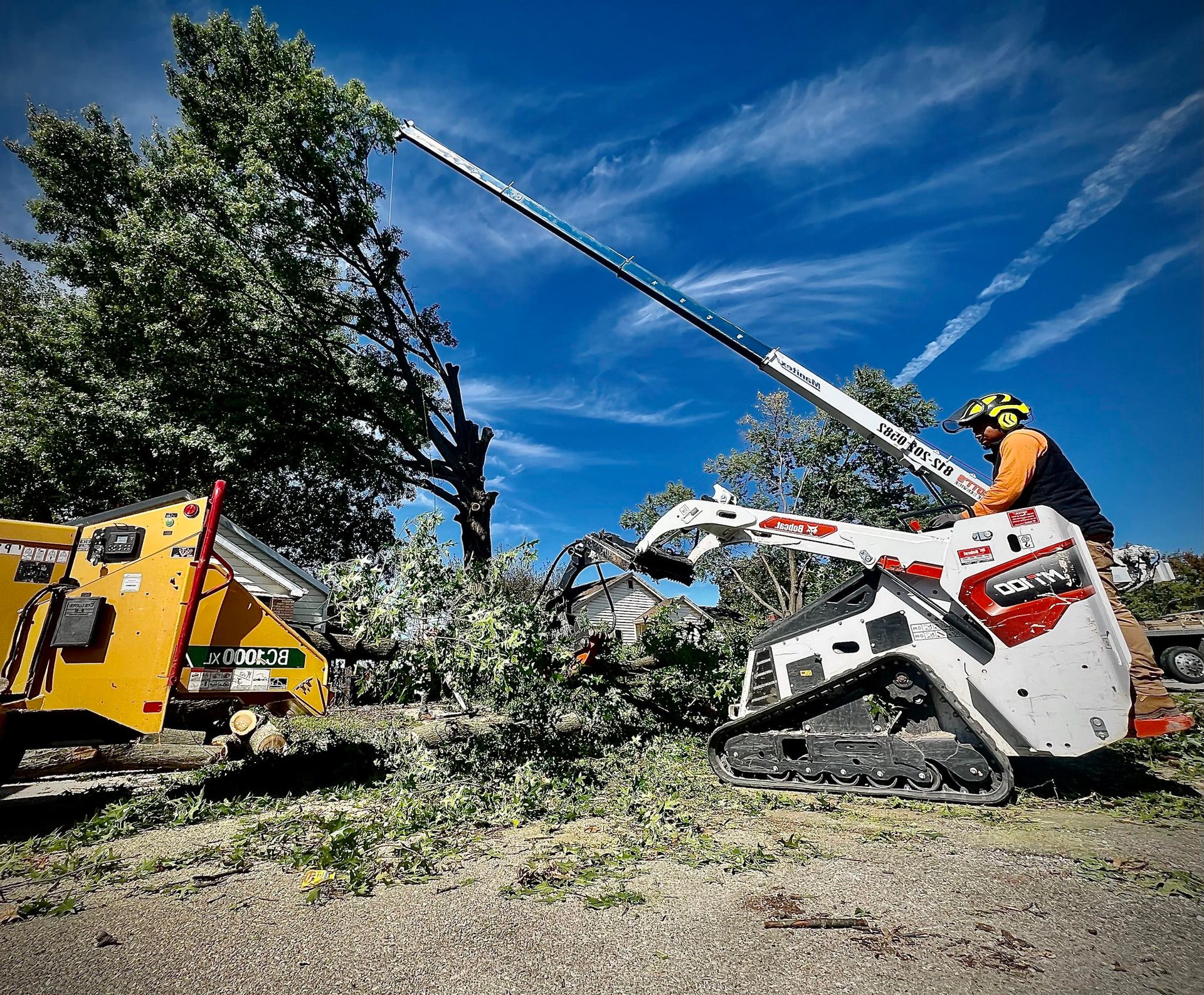 Worker trims tree branches with a white stump grinder or lift beside a yellow chipper under a blue sky.