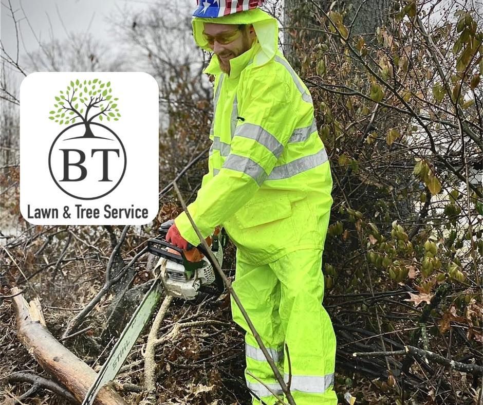 Worker in neon rain gear using a chainsaw amid brush, with BT Lawn & Tree Service logo.