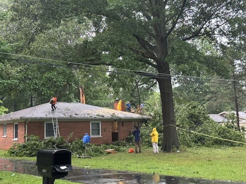 Workers in yellow rain gear clear storm damage near a brick house under a fallen tree.