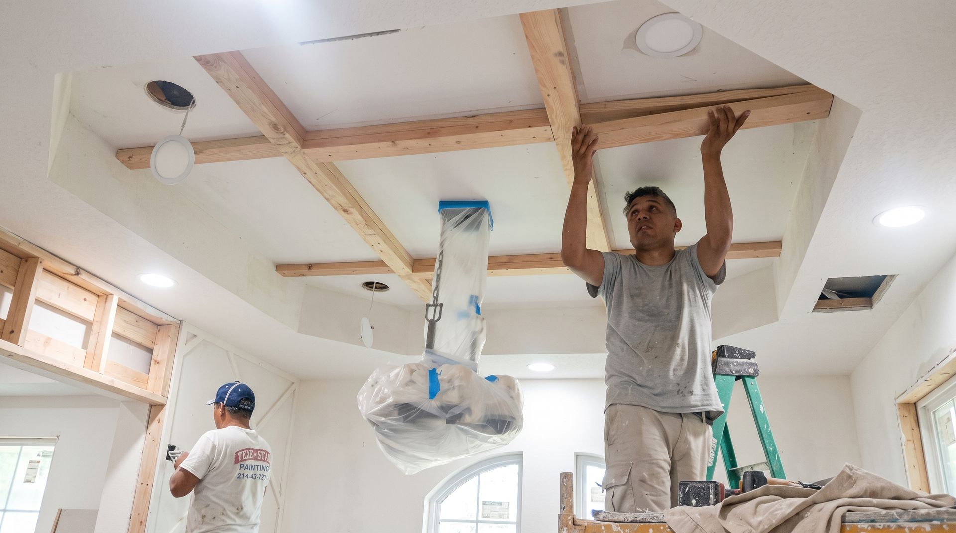 Workers installing drywall on a ceiling in a bright room, with ladders and tools visible.