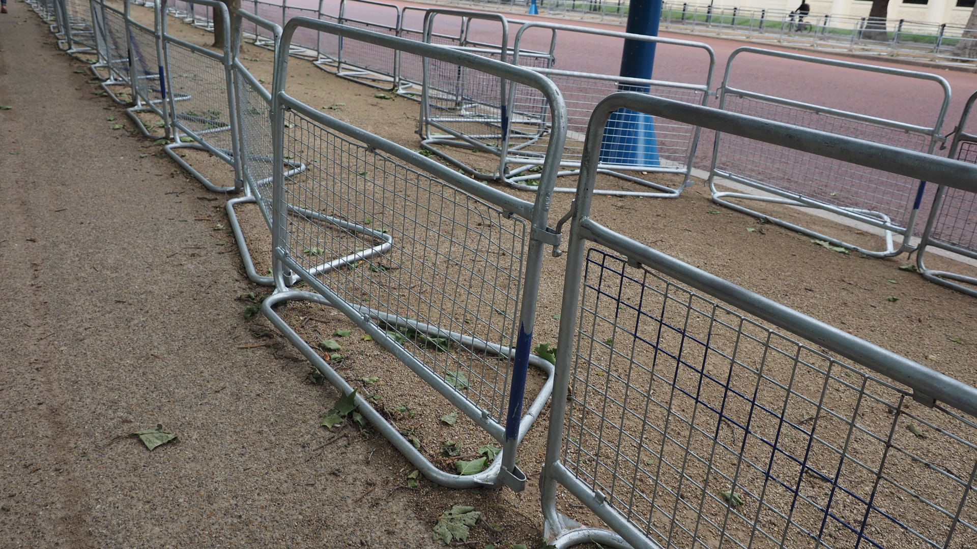Metal barriers along a gravel path beside a running track, with fencing and blue poles in the background