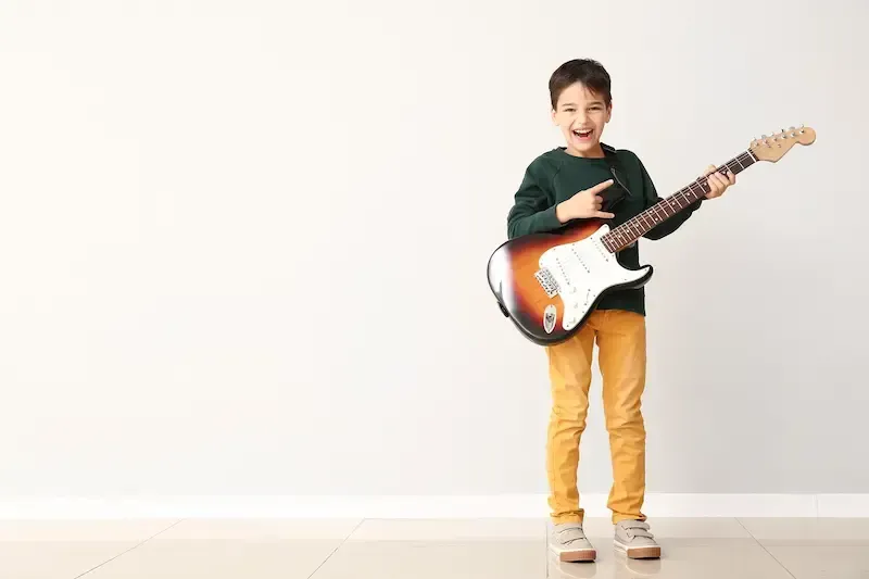 A person in a dark green shirt and yellow pants smiling and holding an electric guitar against a plain white wall.