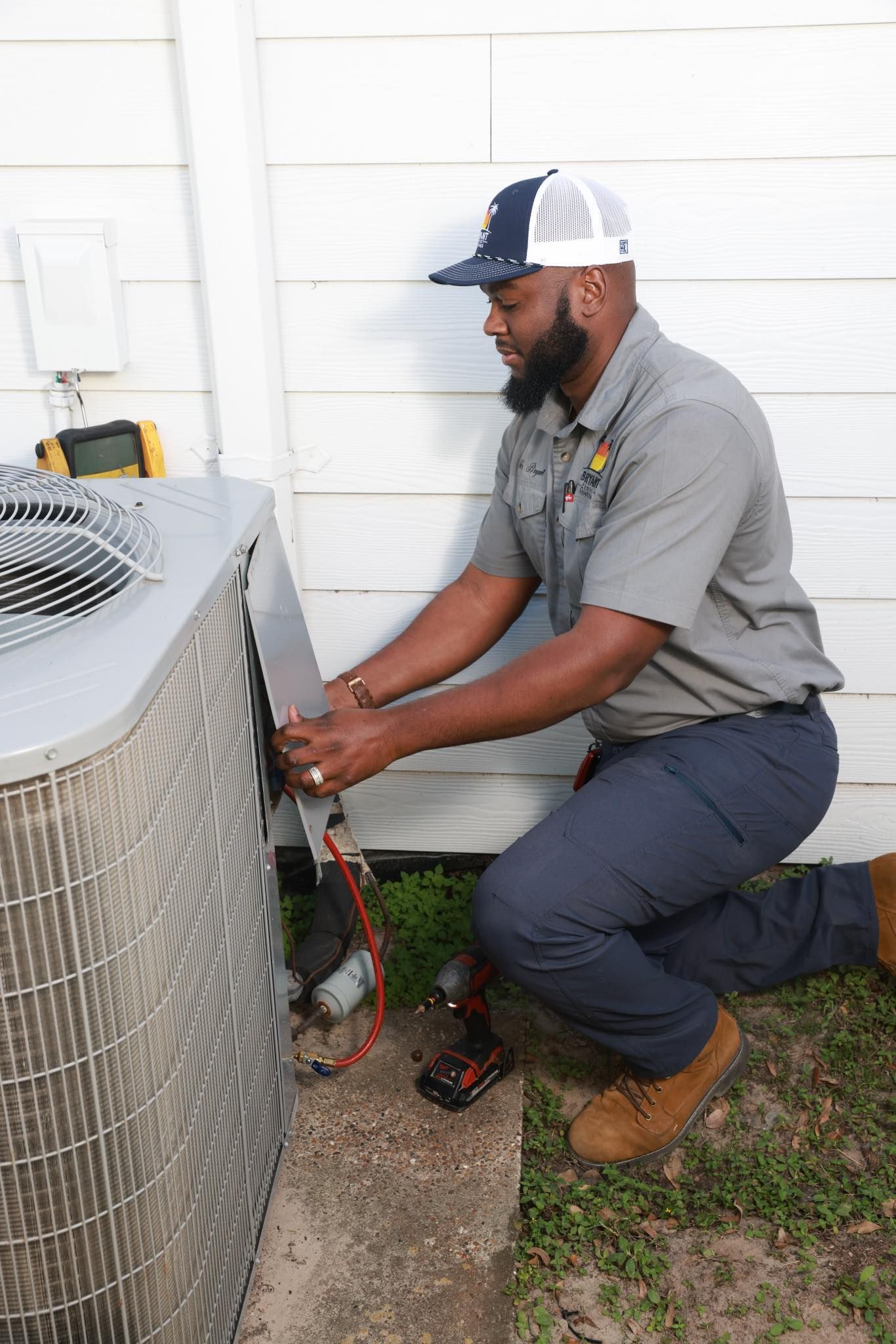 Technician kneeling beside outdoor HVAC unit, adjusting wiring near a white wall.