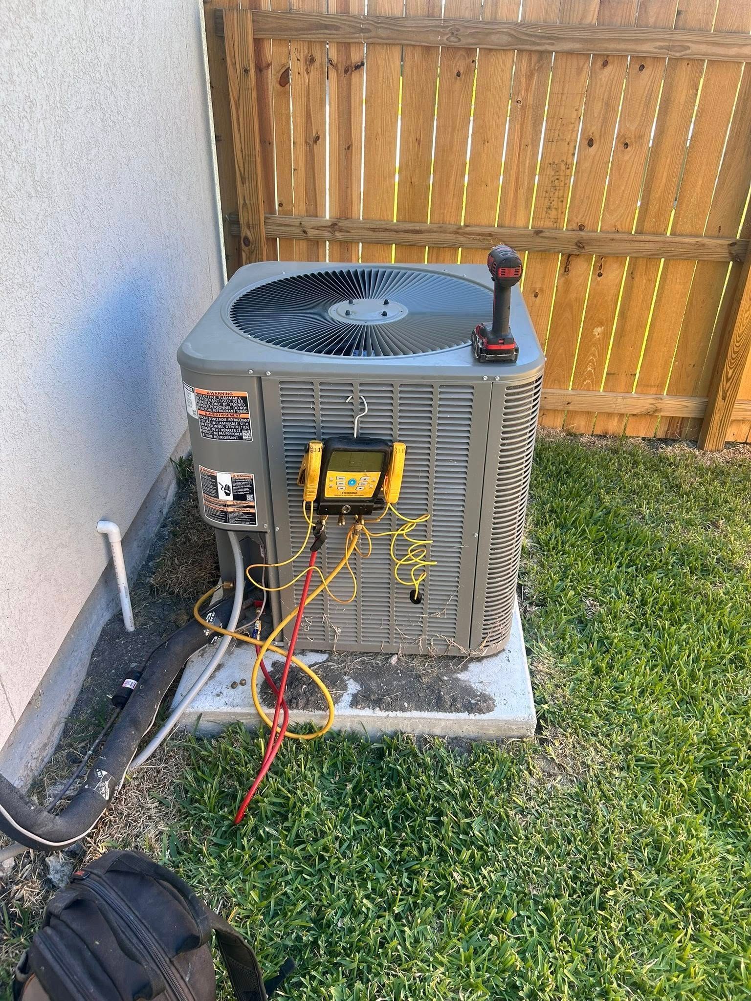 Outdoor air conditioner unit with exposed hoses and wires beside a wooden fence on grass and concrete.
