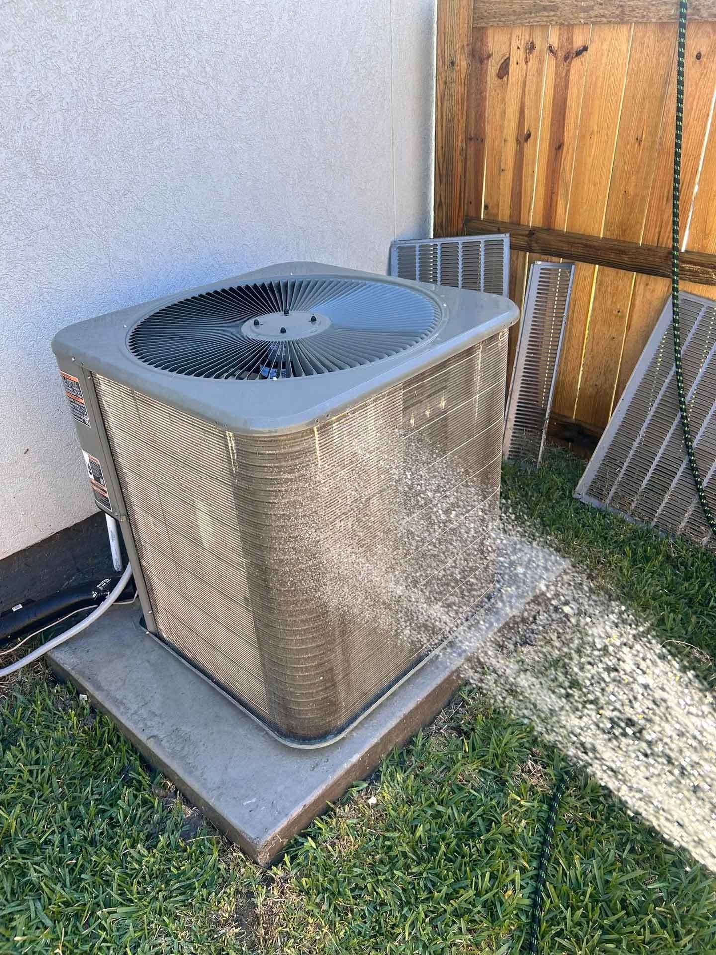 Outdoor HVAC unit spraying water from a hose beside a house and fence