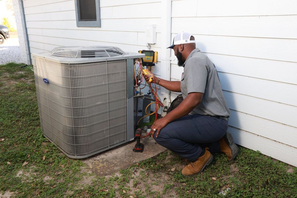Technician kneels beside outdoor HVAC unit, inspecting equipment against a white house wall.