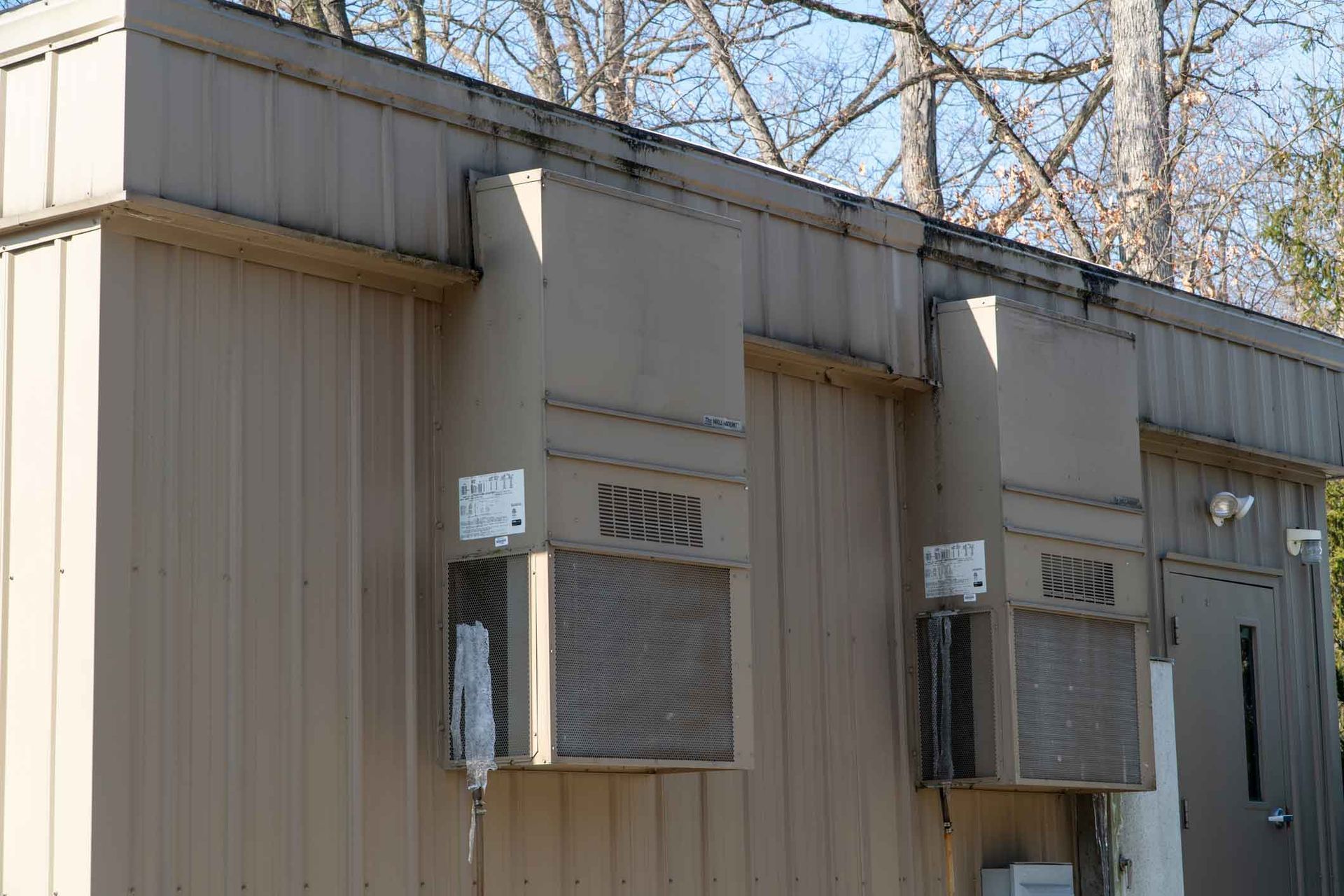 Two beige window air conditioners mounted on a corrugated metal building wall under bare tree branches