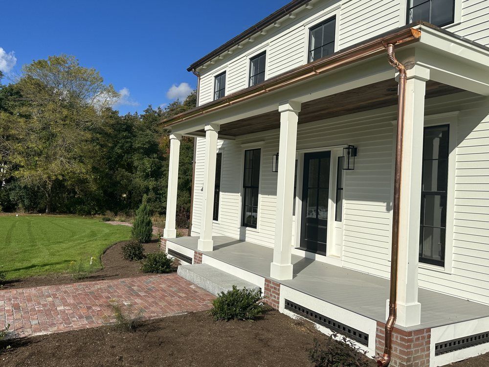 White house porch with columns, black shutters, and a brick walkway beside a green lawn.