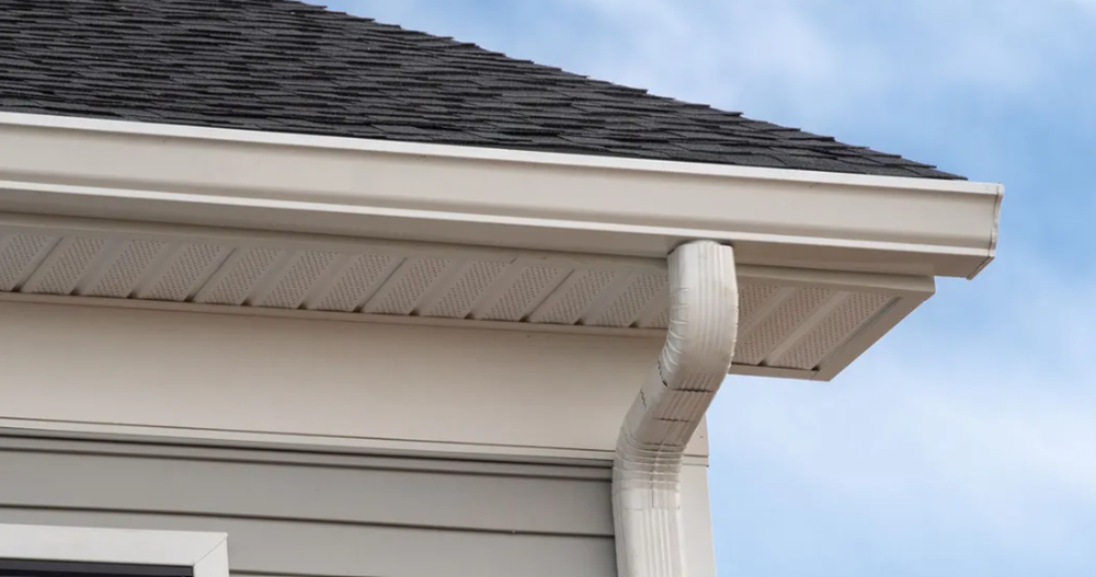 White rain gutter and downspout on a house corner under a shingled roof against a blue sky
