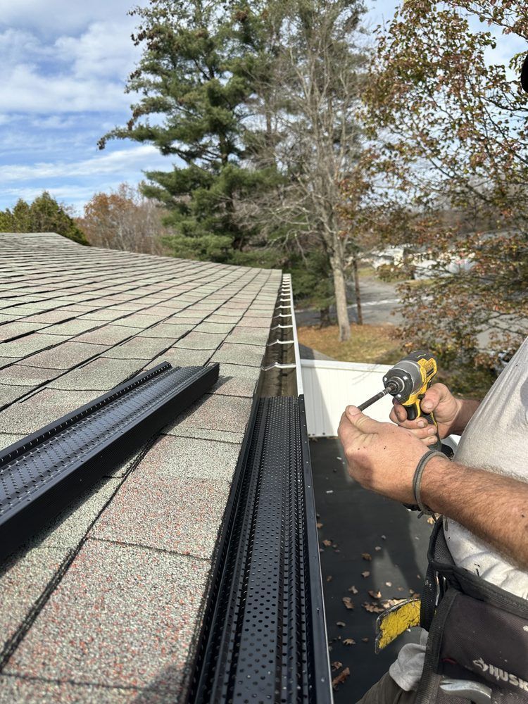 Worker installing black gutter guards on a shingled roof beside trees and a driveway.
