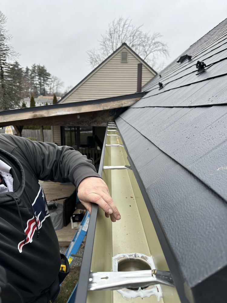 Person installing gutter on a house beside a new roof, holding a tool on a cloudy day