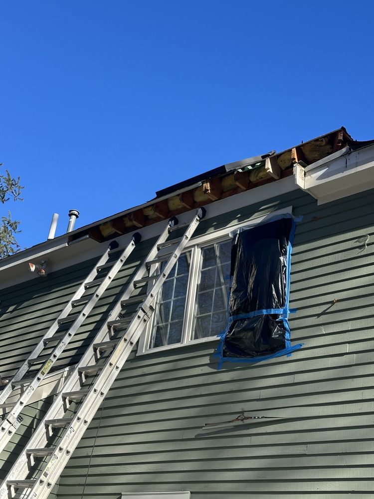 Ladder leaning against house to roof repair, with blue tarp covering an upper window under a clear sky