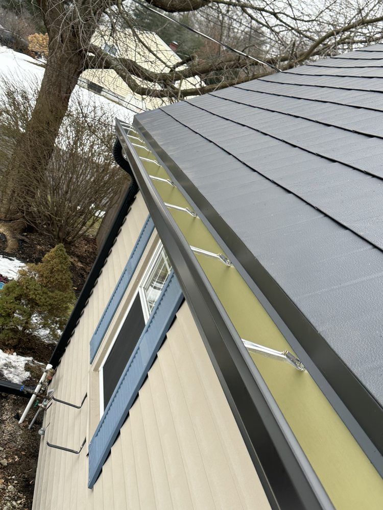 Snow-dusted house roof with gray metal panels and yellow trim beside leafless trees