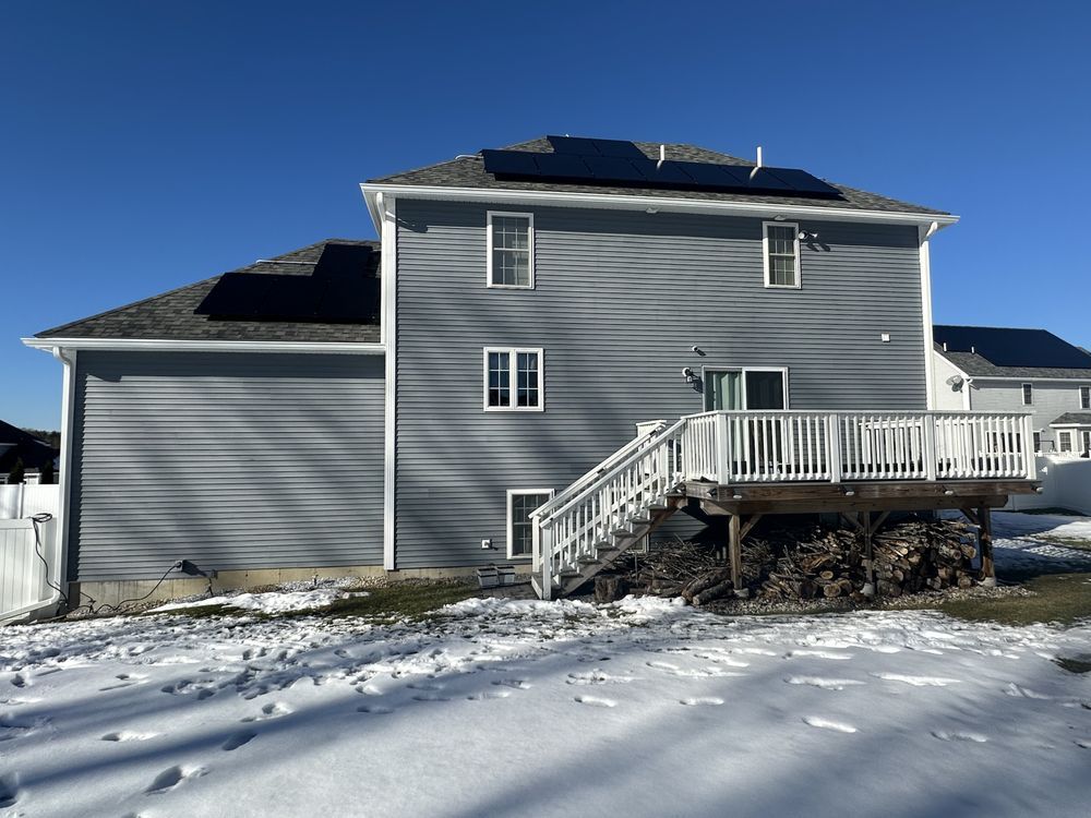 Gray two-story house with a deck and stairs in a snowy yard under a clear blue sky