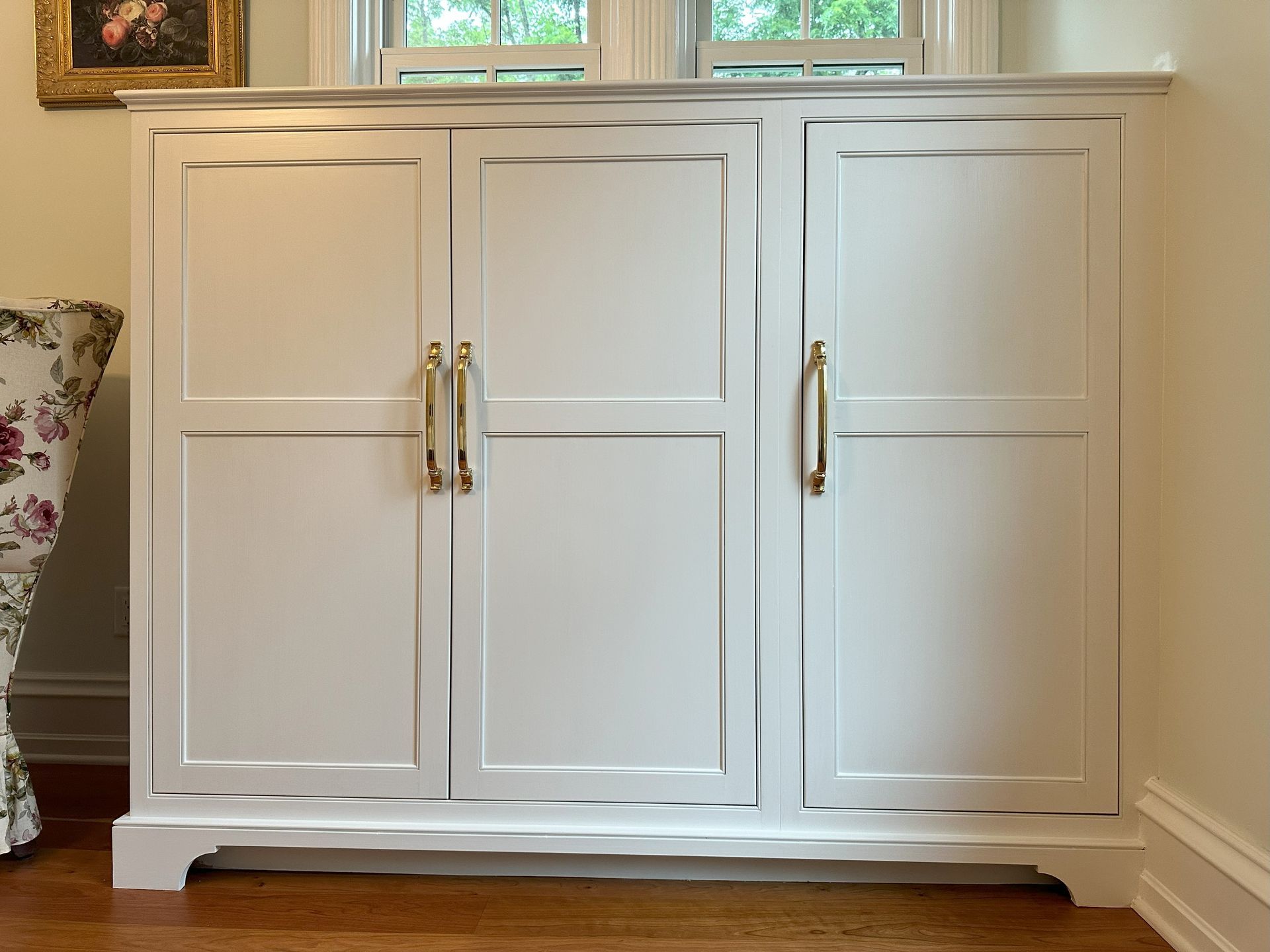 A white cabinet with gold handles is in a room next to a chair.
