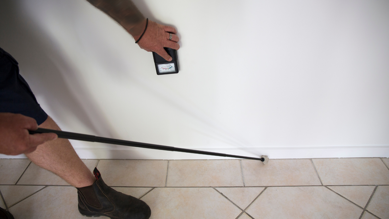 Person using a white cane to feel along a wall while holding a phone in a tiled hallway