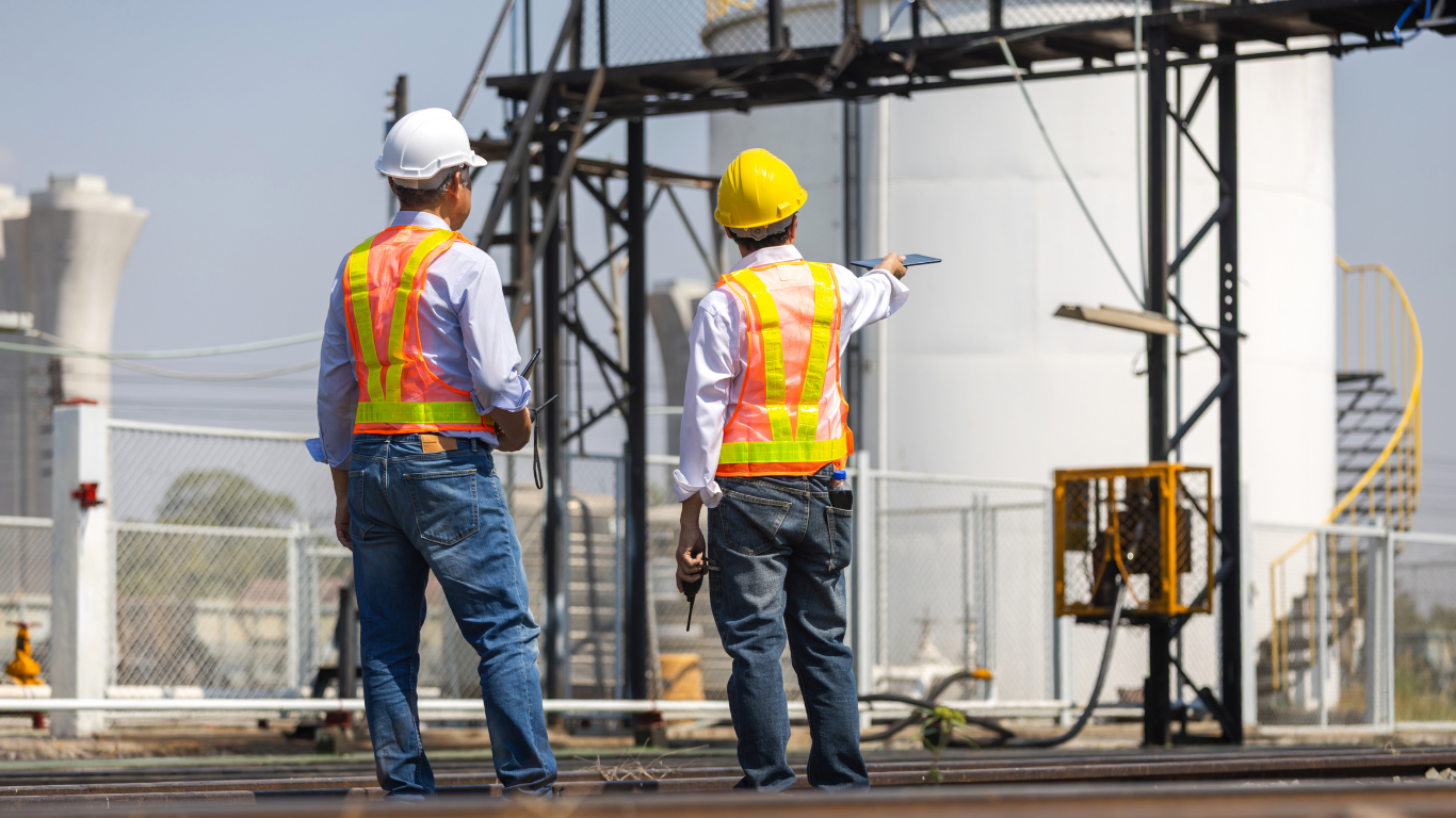 Two construction workers in hard hats and safety vests point at machinery on an industrial site.