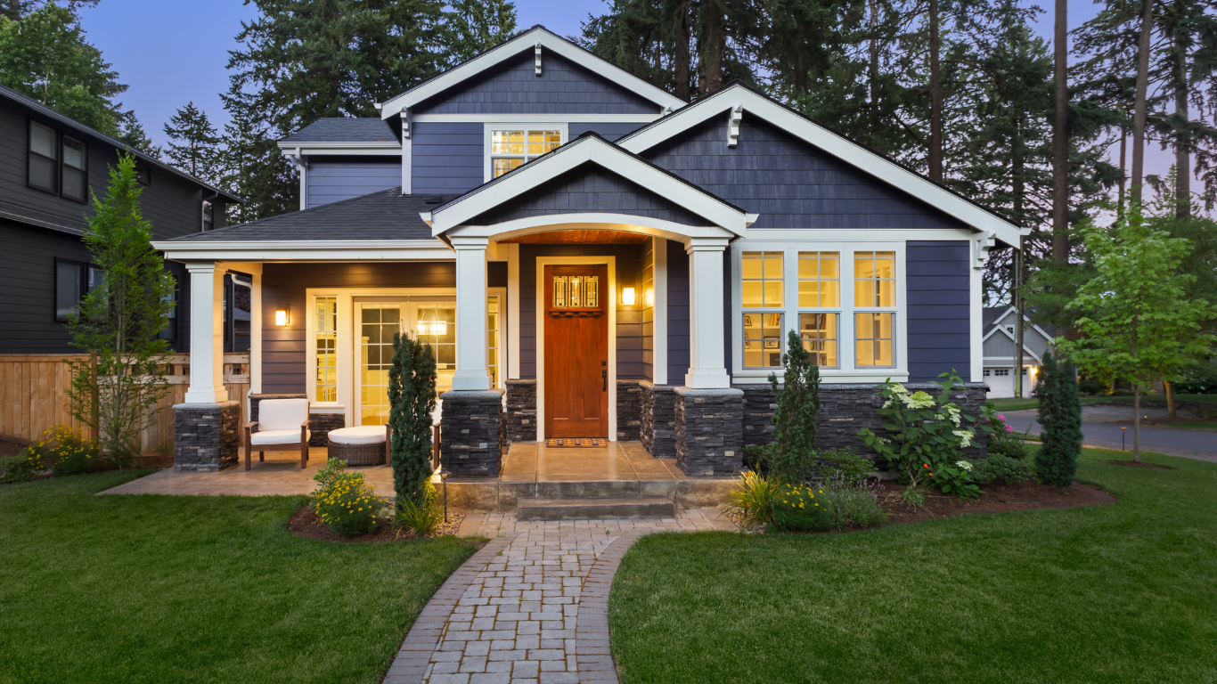 Blue-gray house with warm porch lights, front walkway, and lit windows at dusk