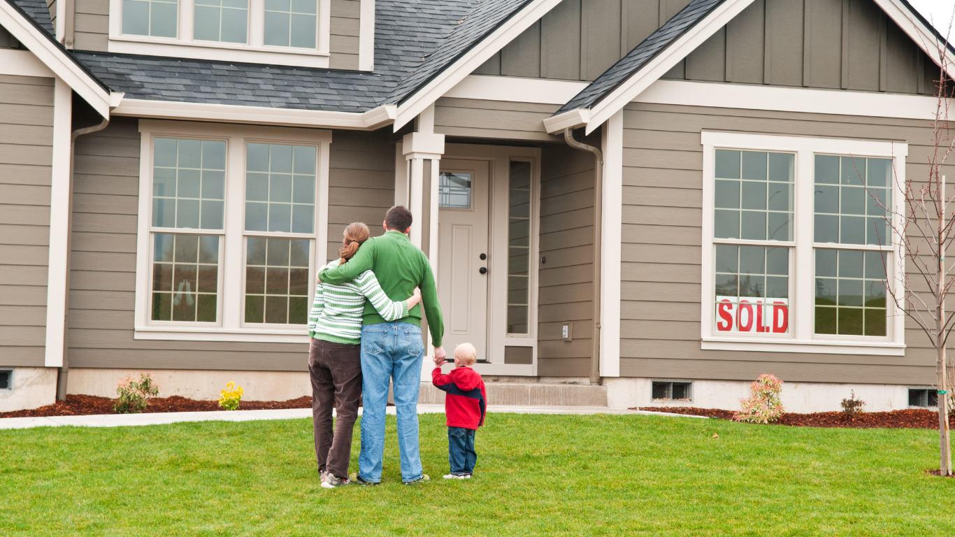 Family standing on lawn in front of a house with a “SOLD” sign in the window.
