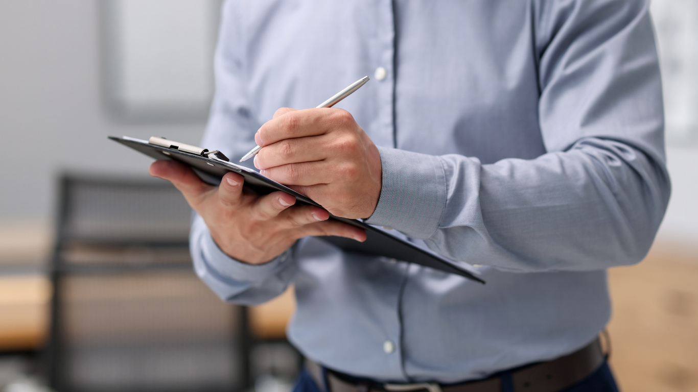 Person in a light blue shirt writing on a clipboard, standing indoors.