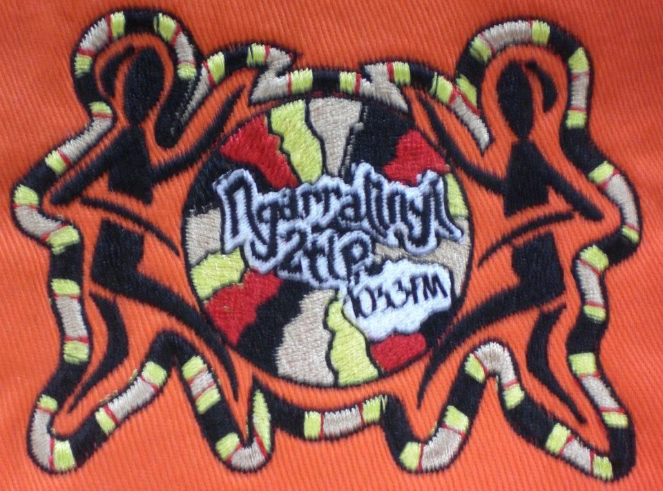 Embroidered patch on orange fabric. Contains stylised figures holding a central design with text, multicoloured, earthy tones — Workwise Clothing Forster in Forster, NSW