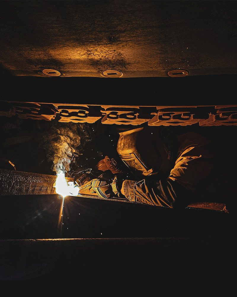 Welder Working Under a Dark Metal Structure, Sparks Flying From the Torch — Workwise Clothing Forster in Taree, NSW