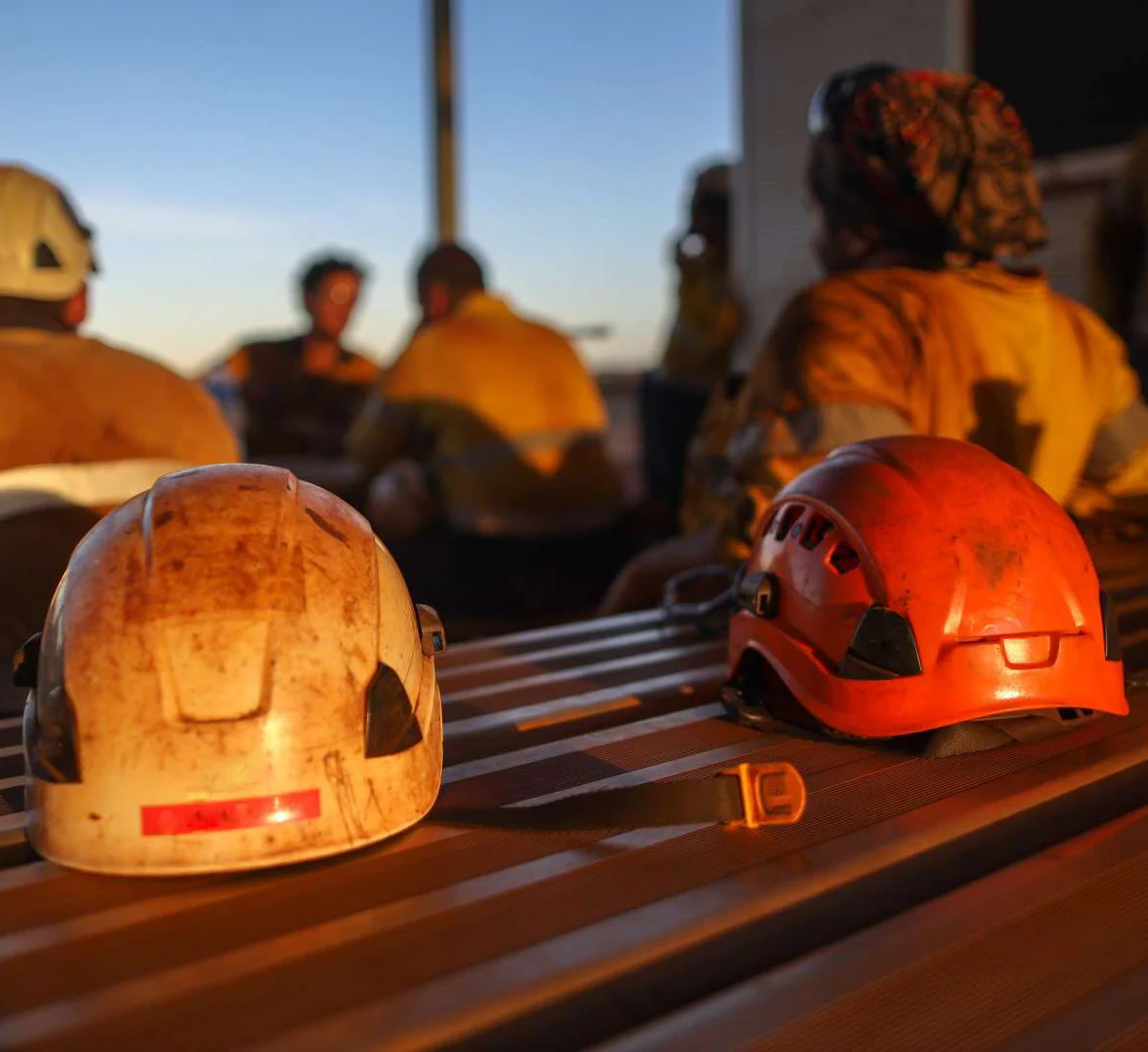 Two dirty work helmets on a table with firefighters in the background during sunset — Workwise Clothing Forster in Forster, NSW