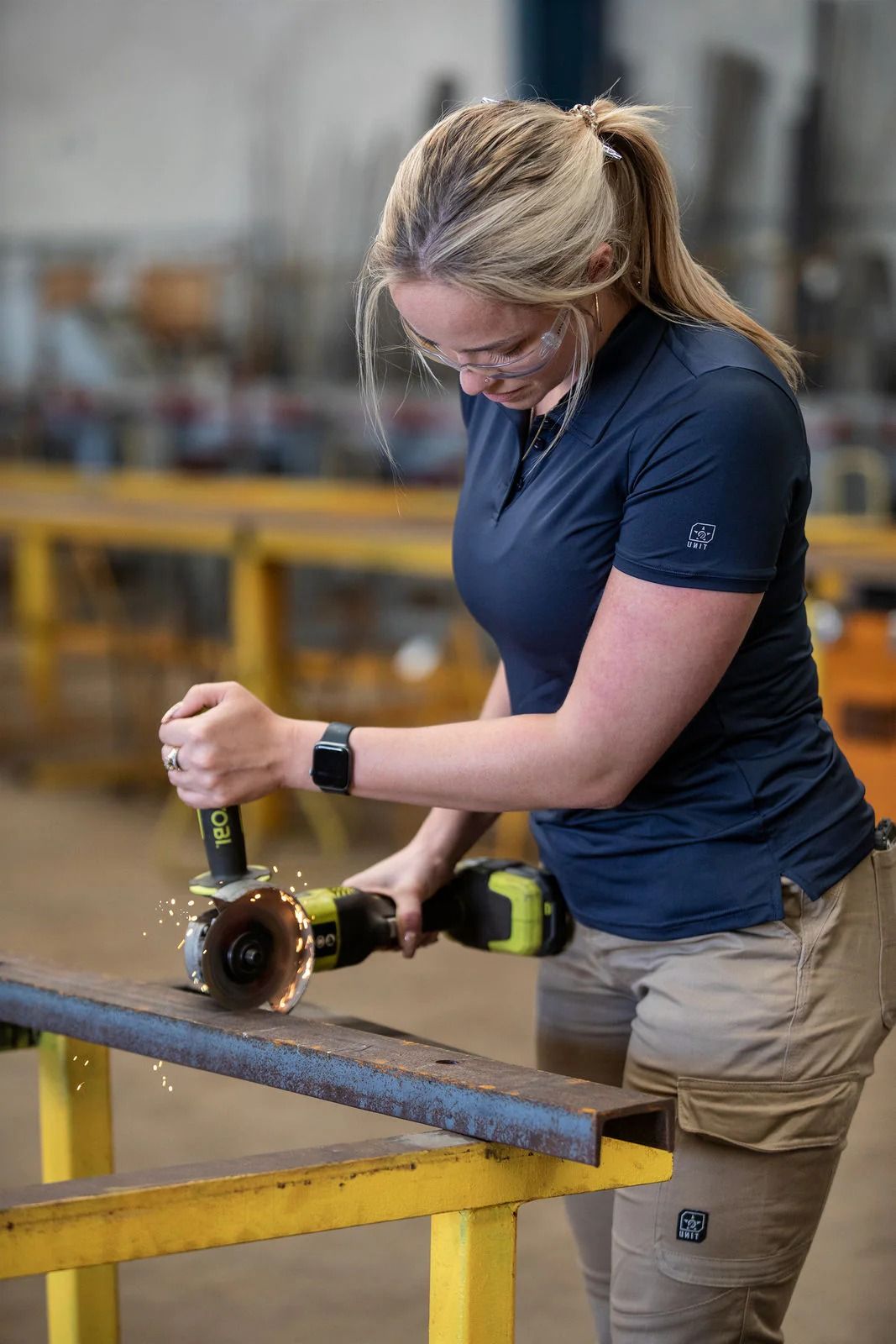 Woman using a grinder on metal in a workshop, wearing safety glasses and work clothes, sparks flying — Workwise Clothing Forster in Forster, NSW