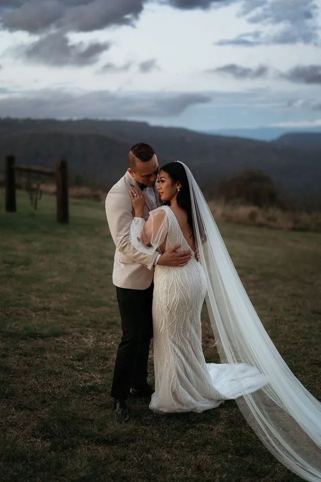 A bride and groom are posing for a picture in a field . the bride is wearing a long veil.