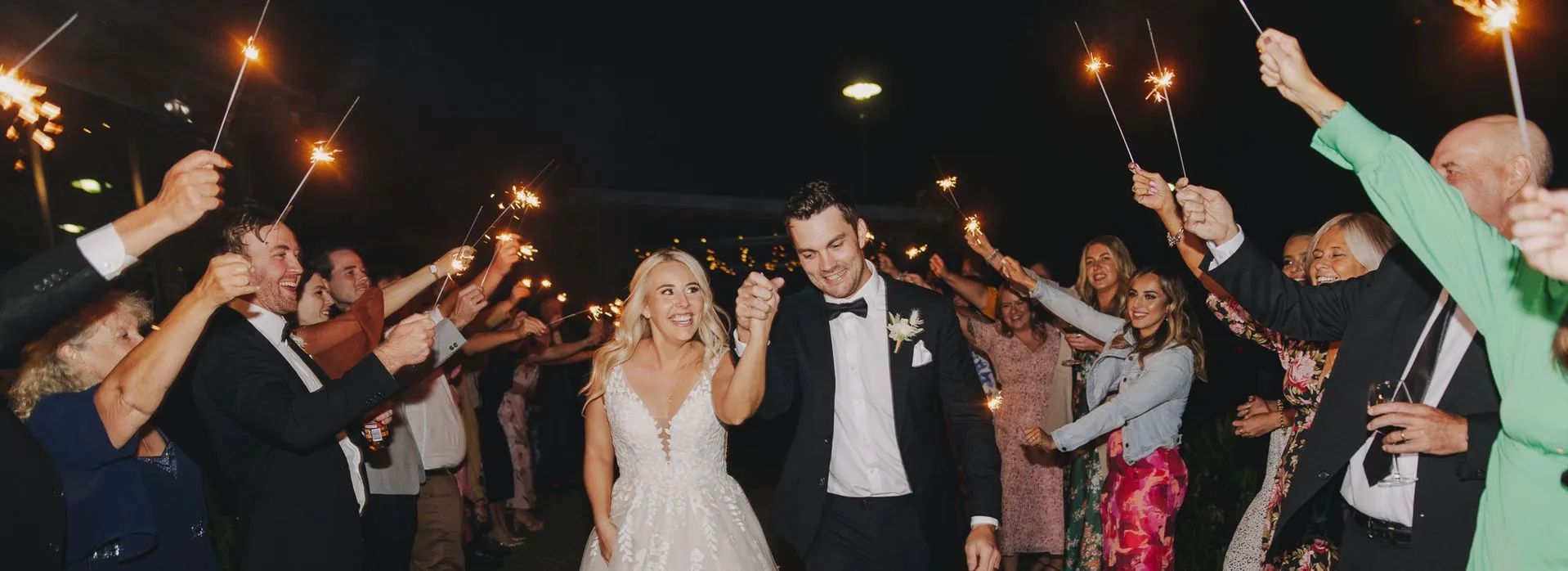 A bride and groom are walking through a crowd of people holding sparklers.