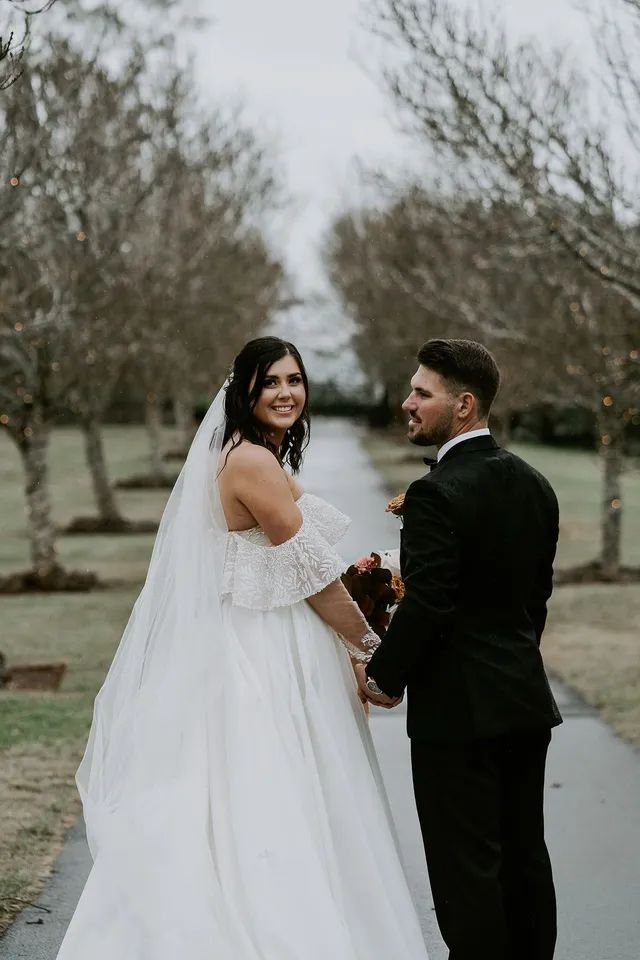 A bride and groom are standing next to each other on a road holding hands.