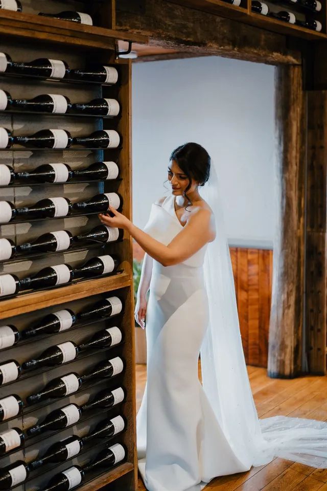 A bride in a white dress is standing in front of a wine rack.