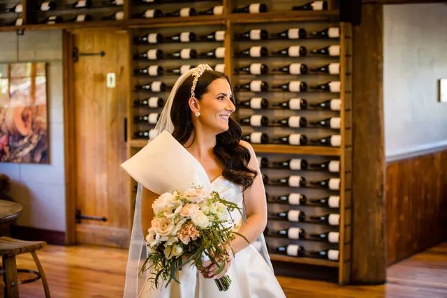A bride is holding a bouquet of flowers in front of a wall of wine bottles.
