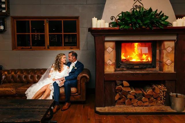 A bride and groom are sitting on a couch in front of a fireplace.