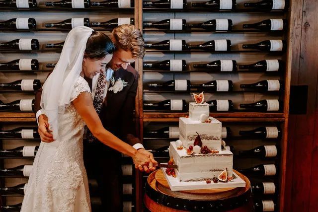 A bride and groom are cutting their wedding cake in front of a wall of wine bottles.