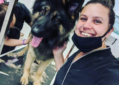 Woman with a mask smiles next to a German Shepherd being groomed; black and tan fur, setting appears to be a pet grooming salon.