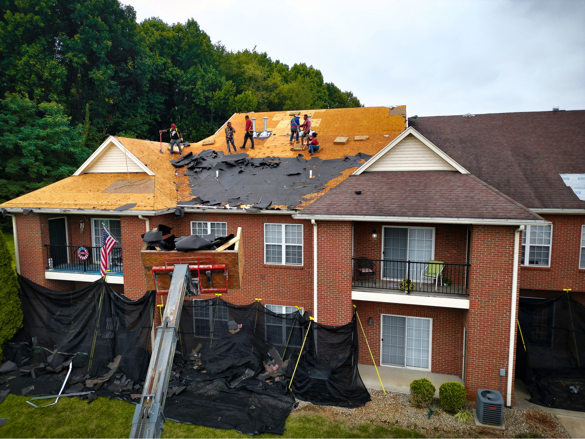 Workers perform repairs on the roof of a multi-story brick apartment building with black protective tarps below.