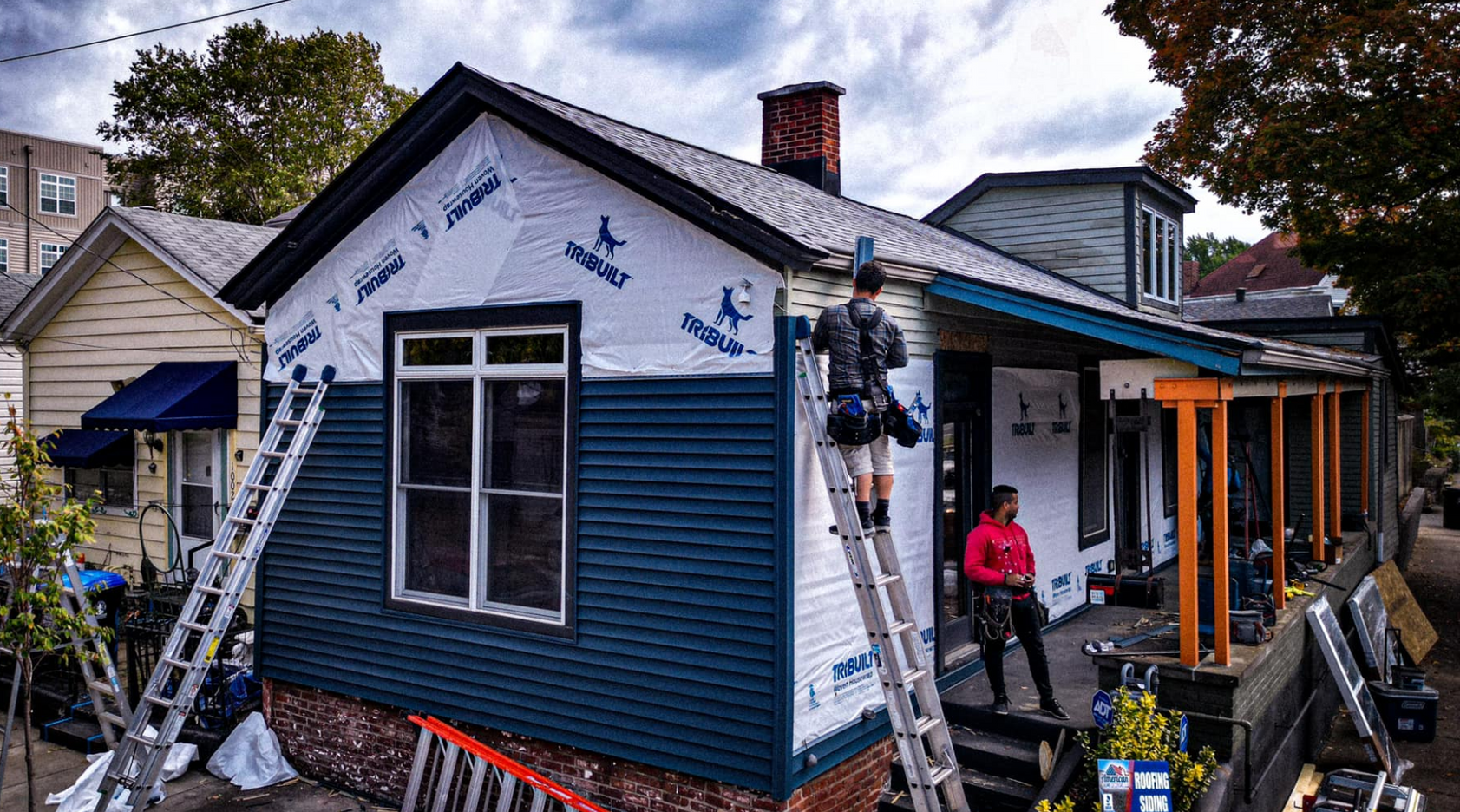 Two construction workers renovate the exterior of a small house, installing blue siding over white protective wrap.