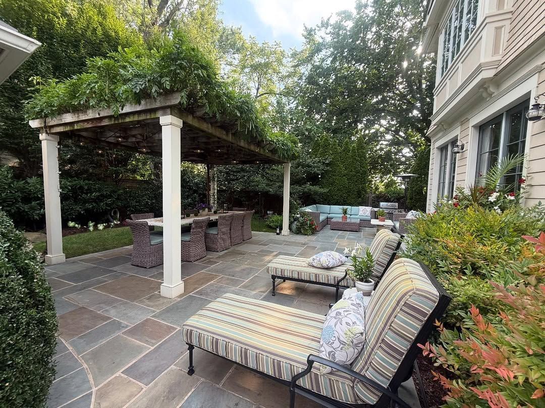 A patio with a dining pergola and lounge chairs next to a house, surrounded by trees and greenery.