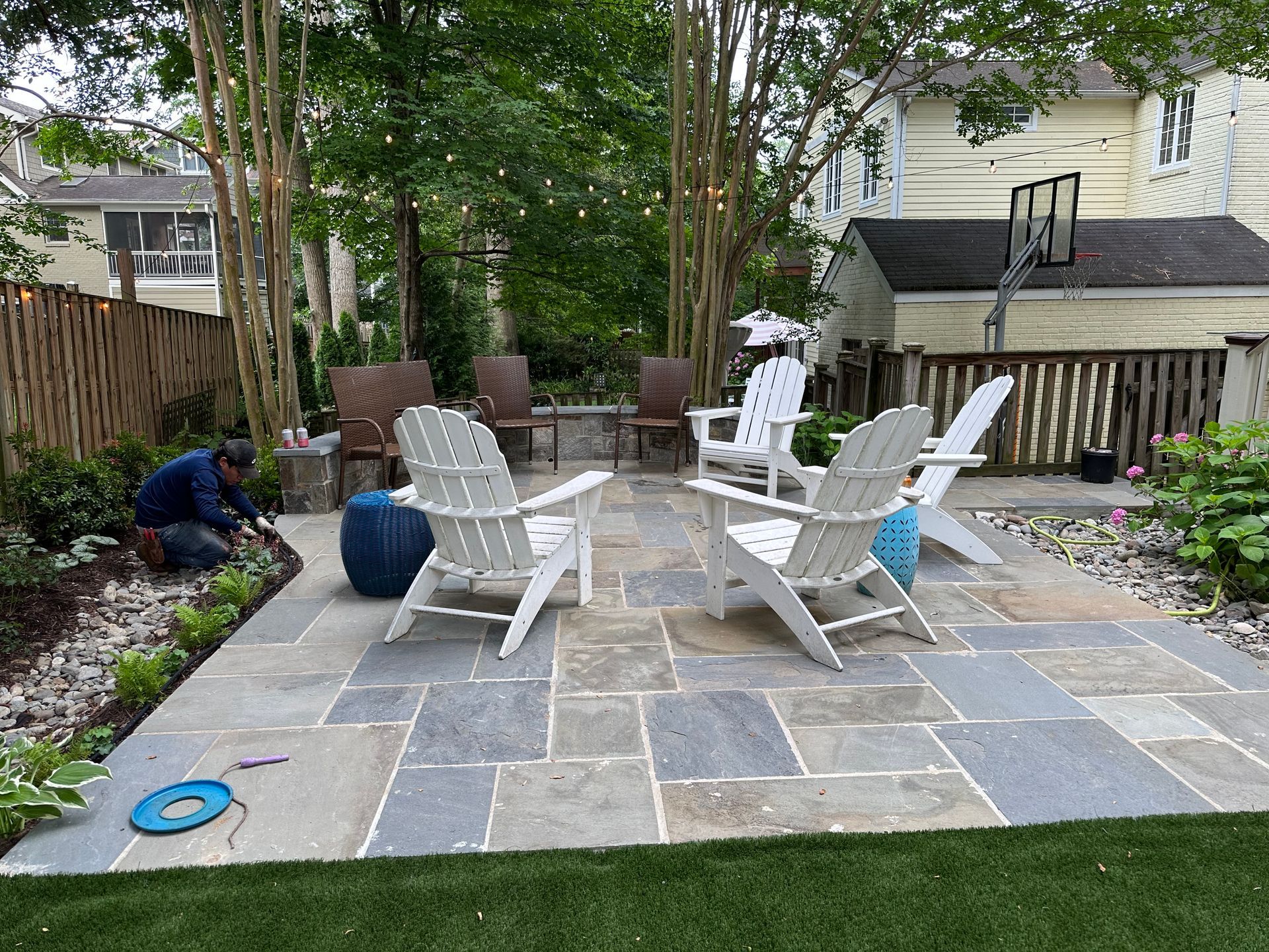 Backyard patio with white chairs and a stone fire pit, surrounded by trees and garden beds