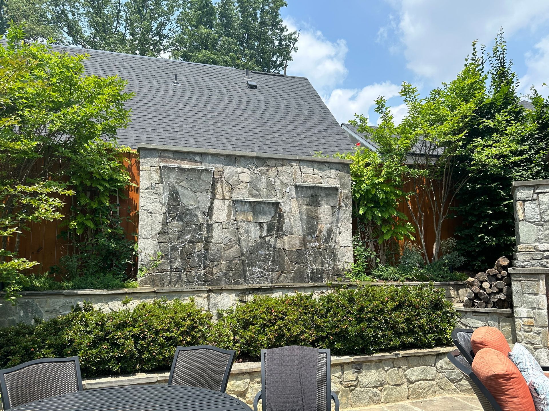 Stone house with slate roof behind a hedge, patio chairs, and lush greenery under a blue sky
