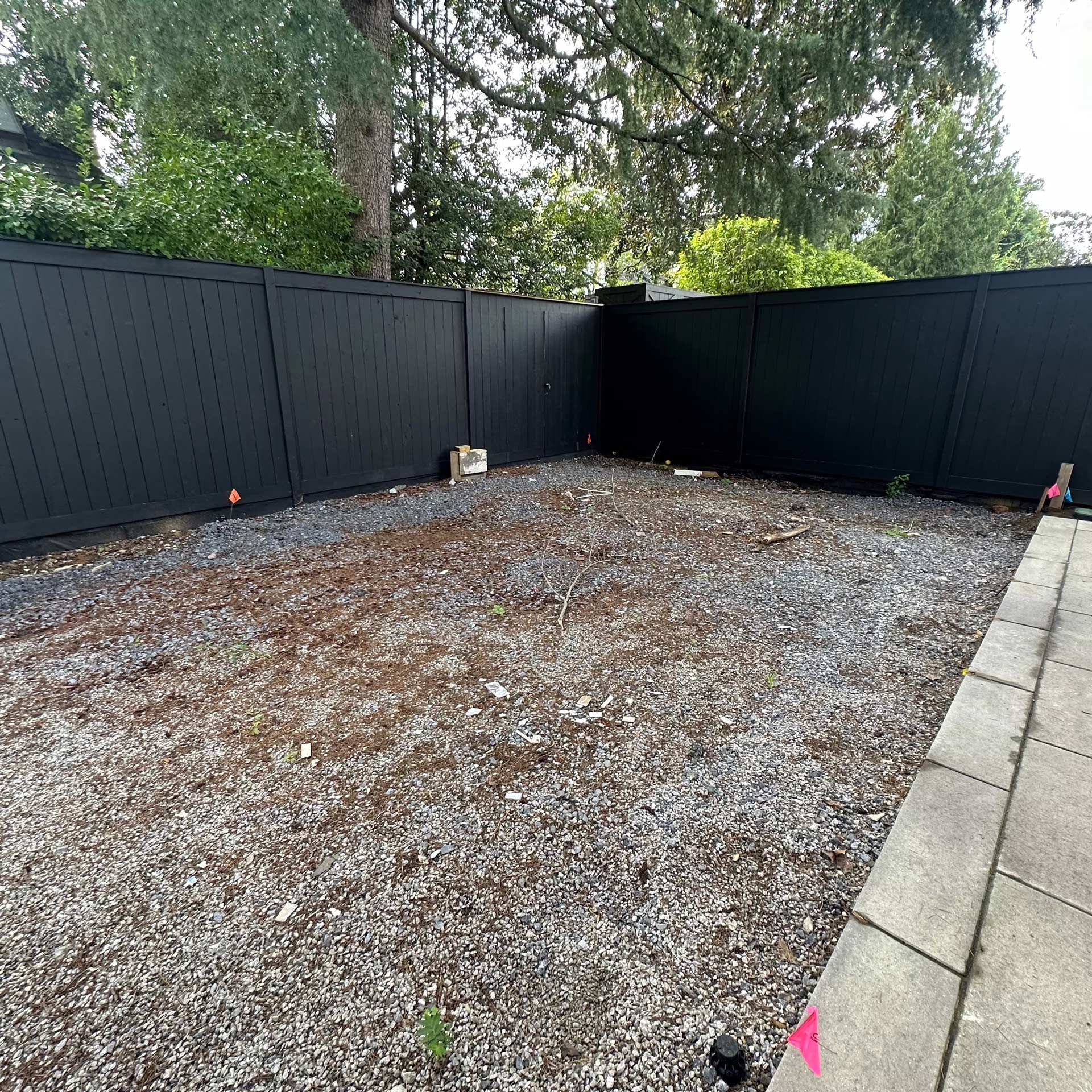 A gravel-covered backyard enclosed by a tall, modern black fence, with a stone patio bordering one side.