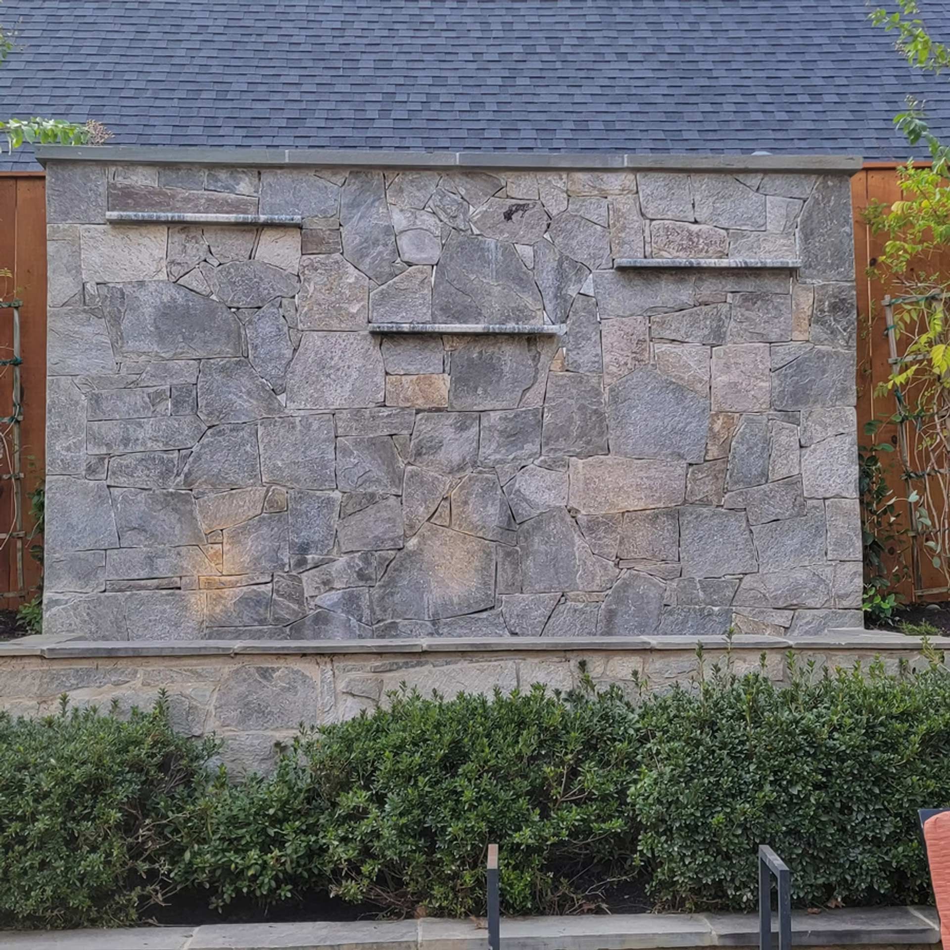 A stone water feature wall with three small protruding ledges, set against wooden fencing and a dark shingled roof.