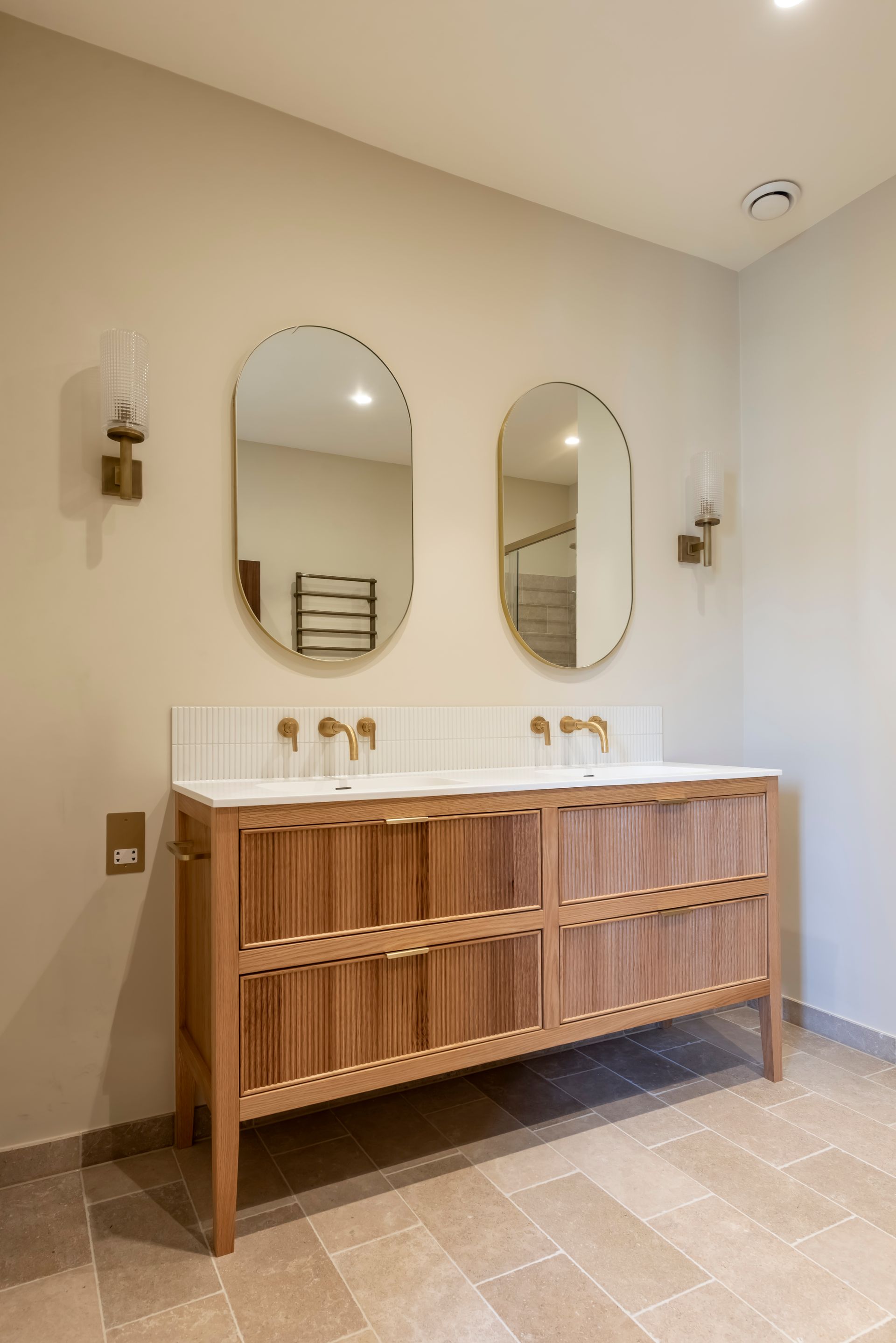 Modern bathroom vanity with twin oval mirrors, wall sconces, and a wooden slatted cabinet on beige tile.
