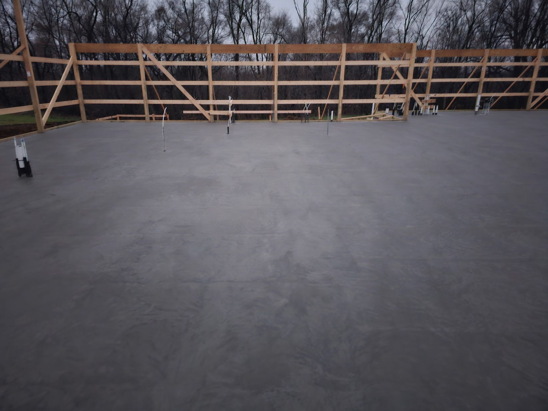 Gray concrete floor with wooden framework against a backdrop of bare trees under an overcast sky.